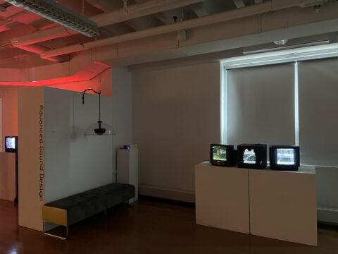 An indoor art exhibition space with a sign on a white partition wall that reads "Advanced Sound Design" vertically. A long, grey padded bench sits in front of the wall under a hanging lamp. To the right, three small, old-fashioned CRT television monitors displaying flickering black-and-white static images are arranged on top of a large white pedestal next to a window with closed blinds.